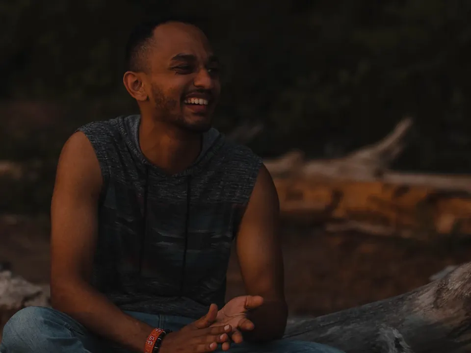A smiling man sits outdoors near driftwood at dusk.