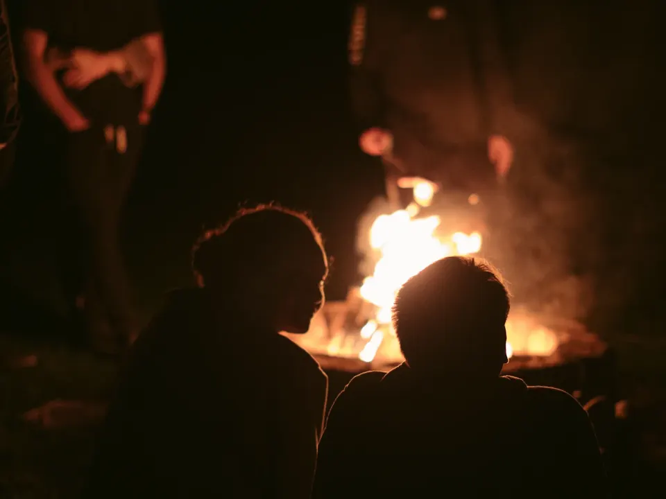 Two people sit by a campfire at night.